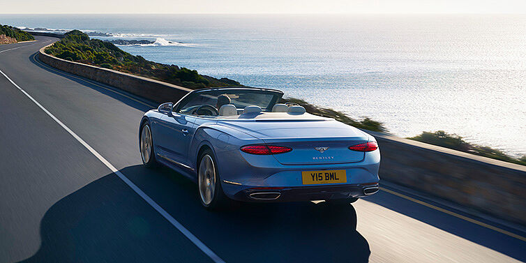 Bentley Maastricht New Bentley Continental GTC Azure convertible rear view in Light Windsor blue paint with 22 inch ten spoke wheels driving dynamically on an ocean road in the sunshine