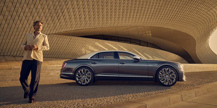 Bentley Maastricht Bentley Flying Spur Mulliner sedan in Rose Gold over Magnetic duotone paint parked by a curved wall with person in suit jacket standing on the left